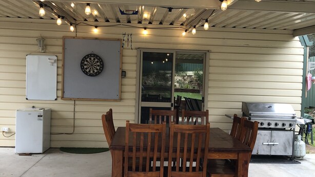 Eine überdachte Terrasse mit Holztisch und -stühlen, einem Dartspiel, einem Kühlschrank und einem Grill.