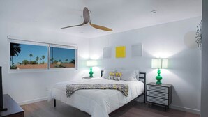 A bedroom with a bed, two bedside tables with lamps, a ceiling fan, and a window with a view of palm trees. - Stylish 2BR+Office, Big Pool, Pickleball and more (Palm Springs)