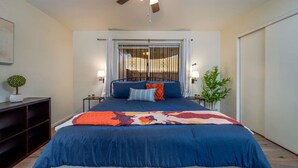 A bedroom with a bed, a ceiling fan, a window with bars, and a nightstand with a lamp.