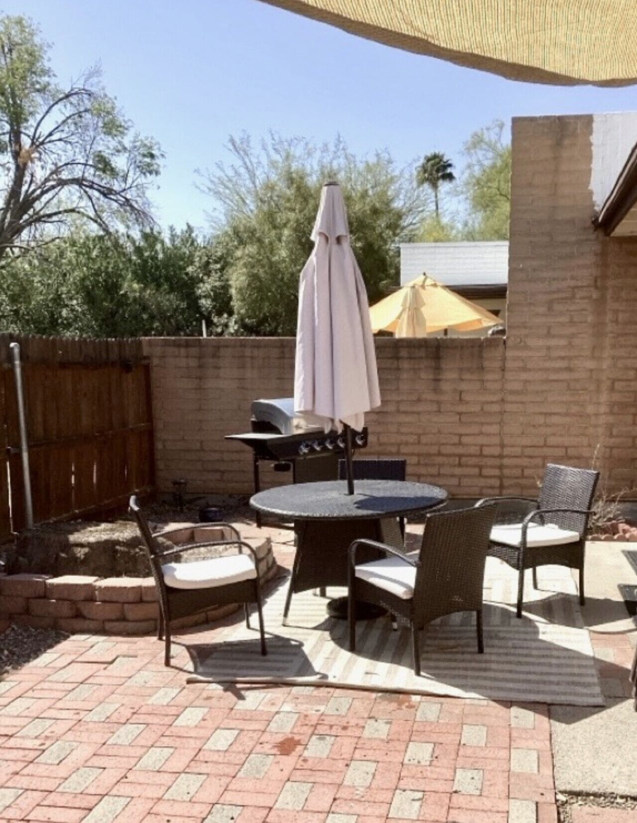An outdoor patio with a brick floor, a round table, four wicker chairs, a barbecue grill, and a large umbrella.