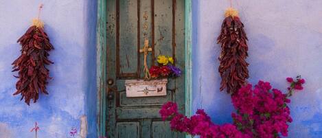 A weathered blue door with a green frame, a small white lantern above, and dried flowers and vibrant pink bougainvillea in front.