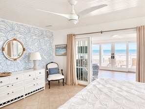 Une chambre à coucher comprenant un lit, une commode, une chaise, un ventilateur de plafond, une grande fenêtre donnant sur la plage et une porte menant à un balcon.