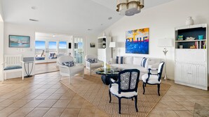 A spacious living room with a large window, a dining area, and a white bookshelf. - 1001 E. Arctic - Out Of The Blue - Unique Outdoor Space- Luxury (Folly Beach)