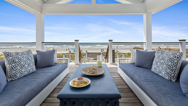 A covered outdoor seating area with a view of the beach, featuring a table with a plate of food and a decorative centerpiece. - 1001 E. Arctic - Out Of The Blue - Unique Outdoor Space- Luxury (Folly Beach)