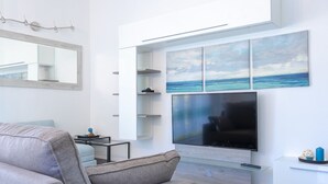 A modern living room with a large flat-screen TV mounted on the wall, a grey sofa, a small coffee table, and a minimalist shelving unit with a painting of the sea. - GIGLIO E MAREA - MAREA (Campo nell'Elba)