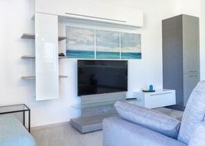 Ein modernes Wohnzimmer mit einem an der Wand befestigten Flachbildfernseher, einem Glastisch, einem grauen Sofa und einer Kommode mit Einbauschränken.