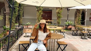 Eine Frau sitzt in einem Straßencafé mit Steinböden, Holztischen und großen Sonnenschirmen.