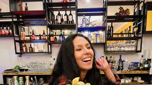 Eine Person hält einen Cocktail mit Limettenscheibe in einer Bar, im Hintergrund Regale voller Flaschen und Gläser.