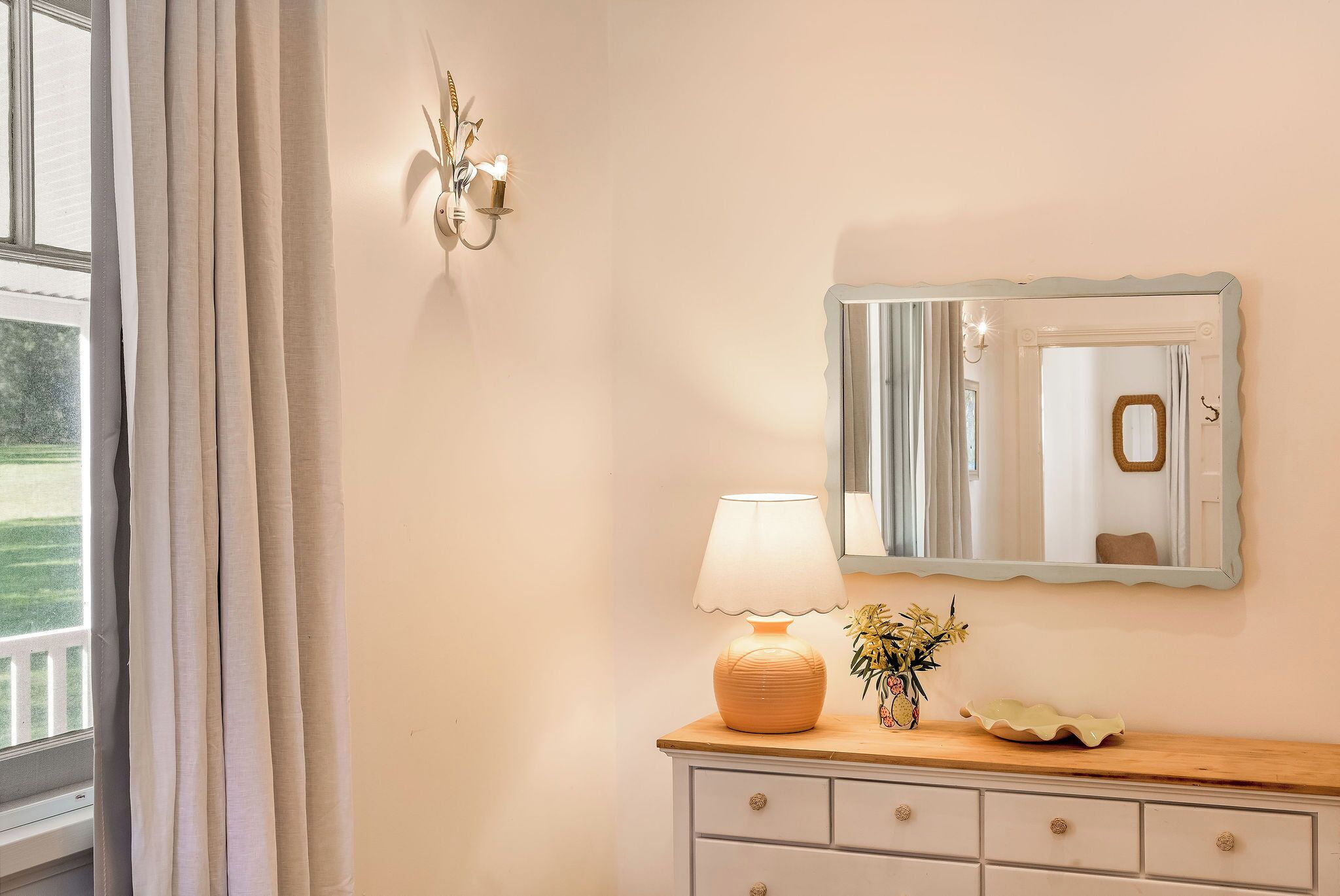 Un baño con tocador, espejo, lámpara y ventana con cortinas.