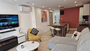A modern living room with a flat-screen TV, a fireplace, a sofa, a yellow chair, and a kitchen area.