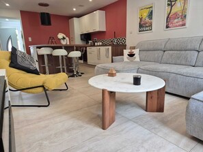 En moderne stue med en grå sofa, en gul lænestol, et rundt sofabord og et køkken i baggrunden.