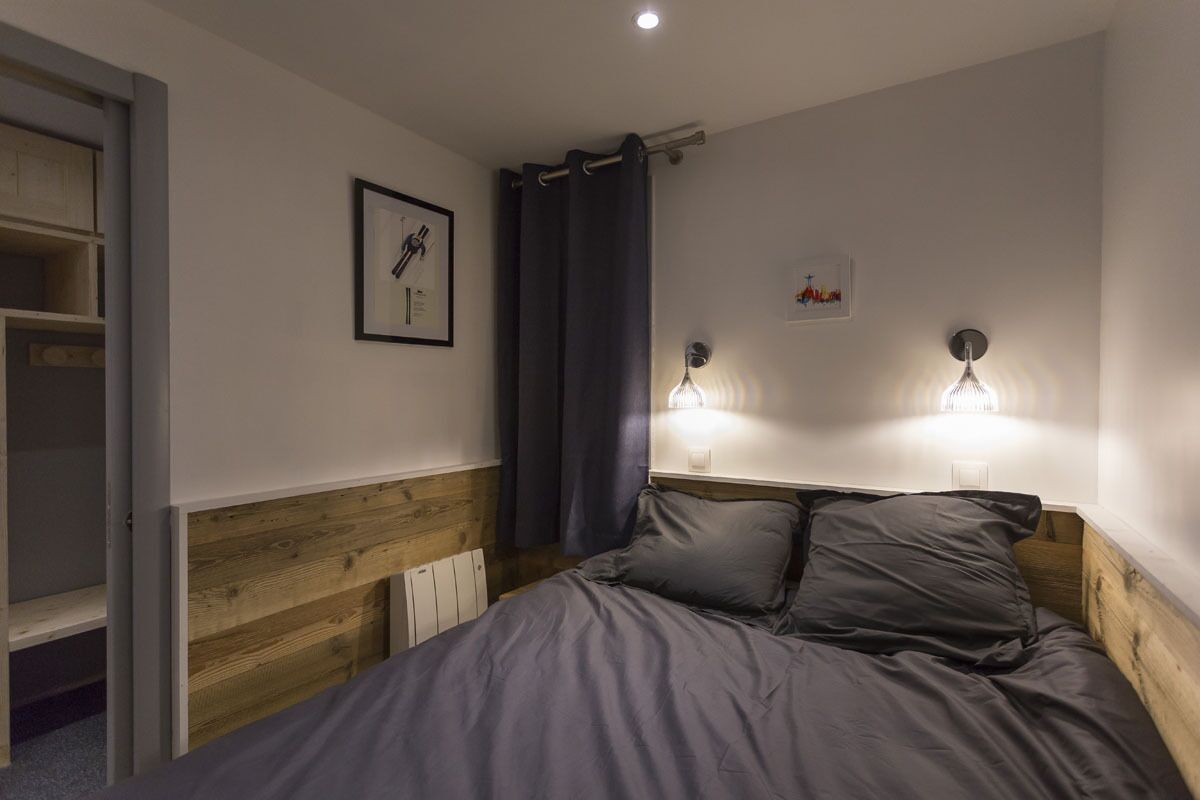 A bedroom with a wooden headboard, a bed with grey bedding, a nightstand with two wall-mounted lamps, a framed picture on the wall, and a partially open door to a closet.