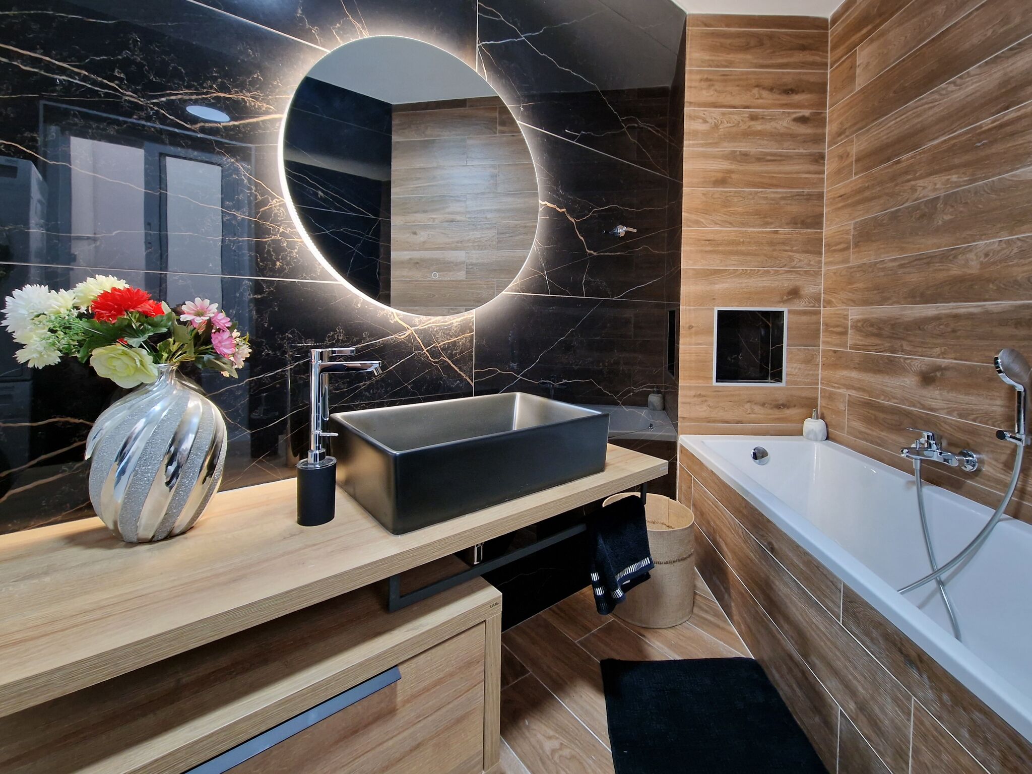 A modern bathroom with a black marble vanity, a rectangular sink, a freestanding bathtub, and a round mirror with a circular light.