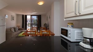 Eine moderne Küche mit weißen Schränken, einer Mikrowelle und einer Kaffeemaschine. Es gibt einen Essbereich mit Tisch und Stühlen, und eine Couch im Hintergrund.