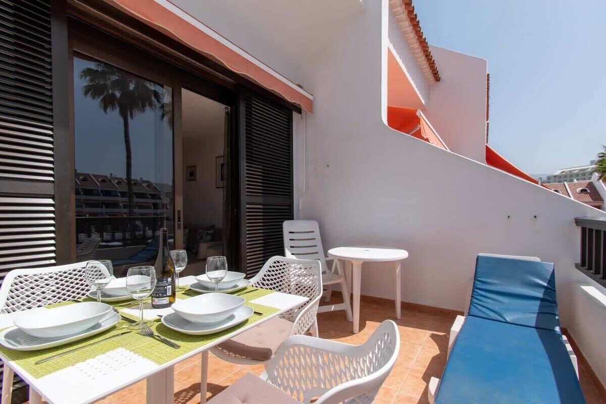 A sunlit outdoor dining area with white plastic chairs and tables, a blue cushioned lounge, and a view of a building with a red roof.