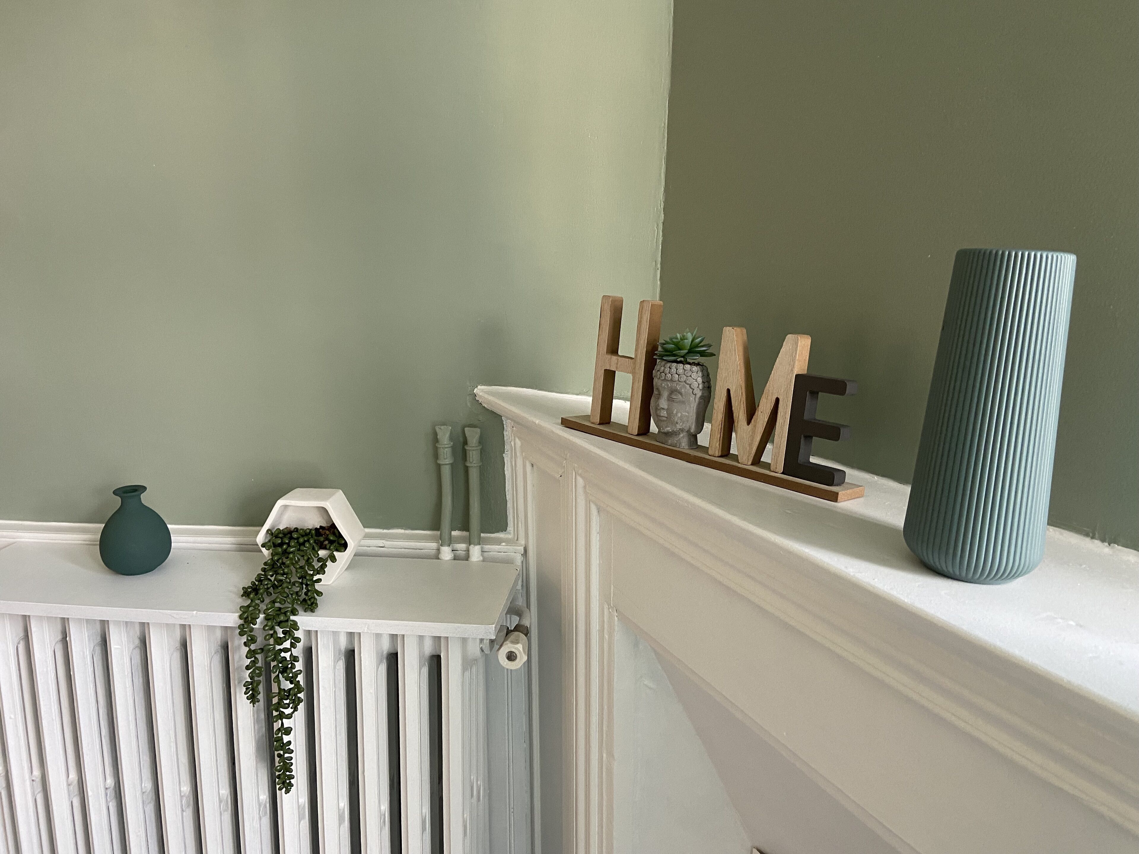 Una repisa de chimenea blanca con un jarrón verde azulado y un letrero de madera que dice 'HOME'.
