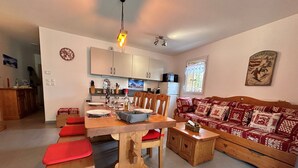 A cozy living room with a wooden dining table, red cushioned benches, a patterned sofa, and a kitchen in the background.