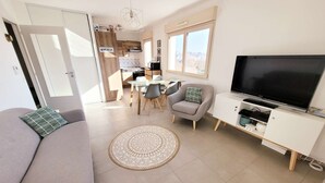 A modern living room with a sofa, a TV, a dining area, and a kitchen in the background.