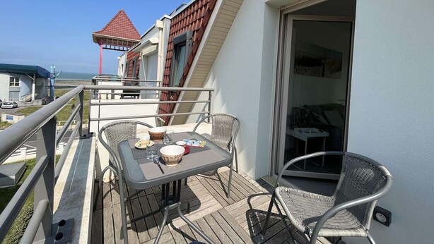 A balcony with a table and chairs, a view of the sea, and a building with a red roof in the background.