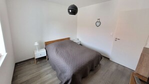 A bedroom with a bed, a nightstand, a lamp, a hanging light fixture, a door, and a remote control on a side table.