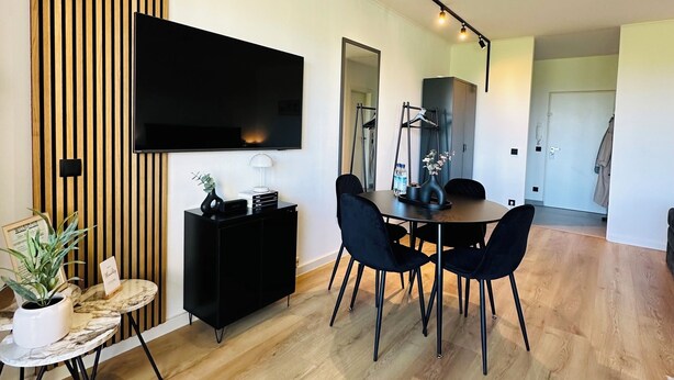A modern living room with a flat-screen TV mounted on the wall, a dining table with chairs, a black cabinet, and a potted plant. - Fewo Haus ,,eckernförde (Schönberg)