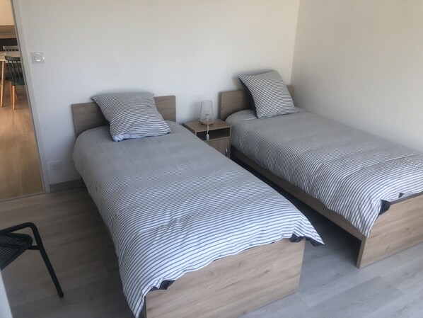 Dos camas individuales con ropa de cama a rayas, cabeceros de madera, una mesita de noche con lámpara y una silla.