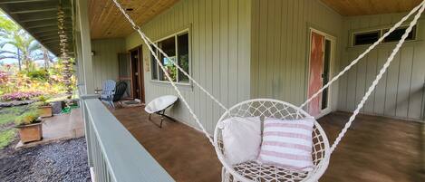 A covered porch with a white hammock chair, a wooden ceiling, and a view of a garden.