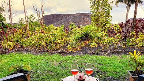 A patio with two chairs and a table, two glasses of red drink, and a lush garden with various plants and a mountain in the background. - Lava House (Pahoa)