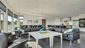 A modern living room with a white coffee table, black leather sofas, and a kitchen area with white cabinets and a black oven. - 4 bedroom nice home in Ebeltoft (Ebeltoft)