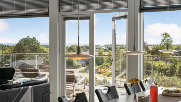 A modern dining area with a panoramic view, featuring a table set with chairs, a vase with flowers, and a glass of red drink. - 4 bedroom nice home in Ebeltoft (Ebeltoft)