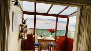 Une pièce avec une grande fenêtre, une table entourée de chaises, un manteau de fourrure accroché et un lit recouvert d’une couverture à motifs bleus.
