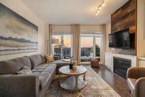 A modern living room with a sectional sofa, a round coffee table, a fireplace, and a large TV mounted on a wooden wall. - Sunrises Near DT & Stampede 2 BR 2 BA w AC (Calgary)