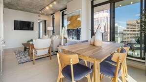 Un salon moderne avec une table à manger en bois, des sièges confortables, une grande peinture abstraite et une vue sur la ville à travers de grandes fenêtres.