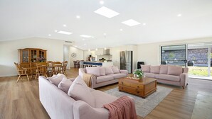 A spacious living room with two sofas, a dining area with a wooden table and chairs, a kitchen with modern appliances, and a large sliding glass door leading to a garden. - The Hideaway at Sutton Forest Retreat (Sutton Forest)