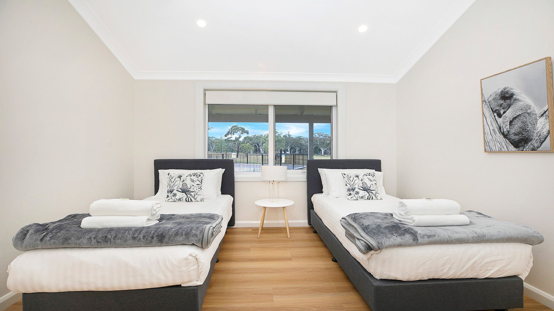 A modern bedroom with two beds, a small table, and a large window offering a view of trees and a fence.
