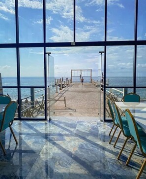 Vista de dentro de um edifício com grandes janelas, com vista para um cais e o oceano.