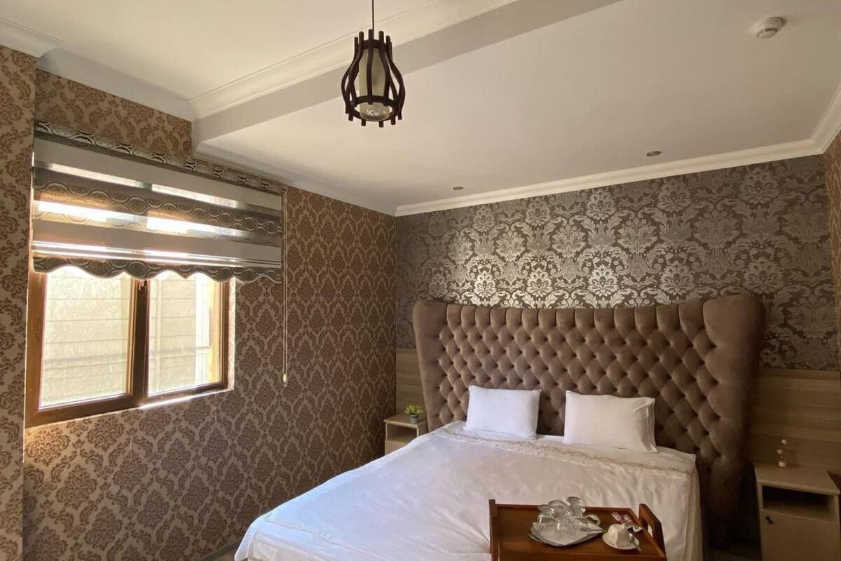 A bedroom with a large bed, a chandelier, patterned wallpaper, and a window with blinds.