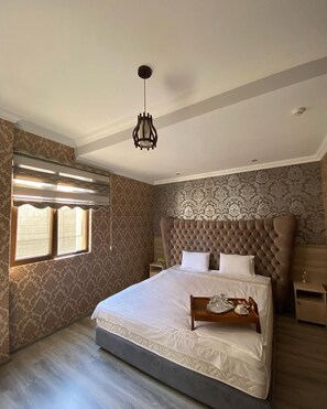 Quarto com cama grande, lustre, papel de parede estampado e janela com persianas.