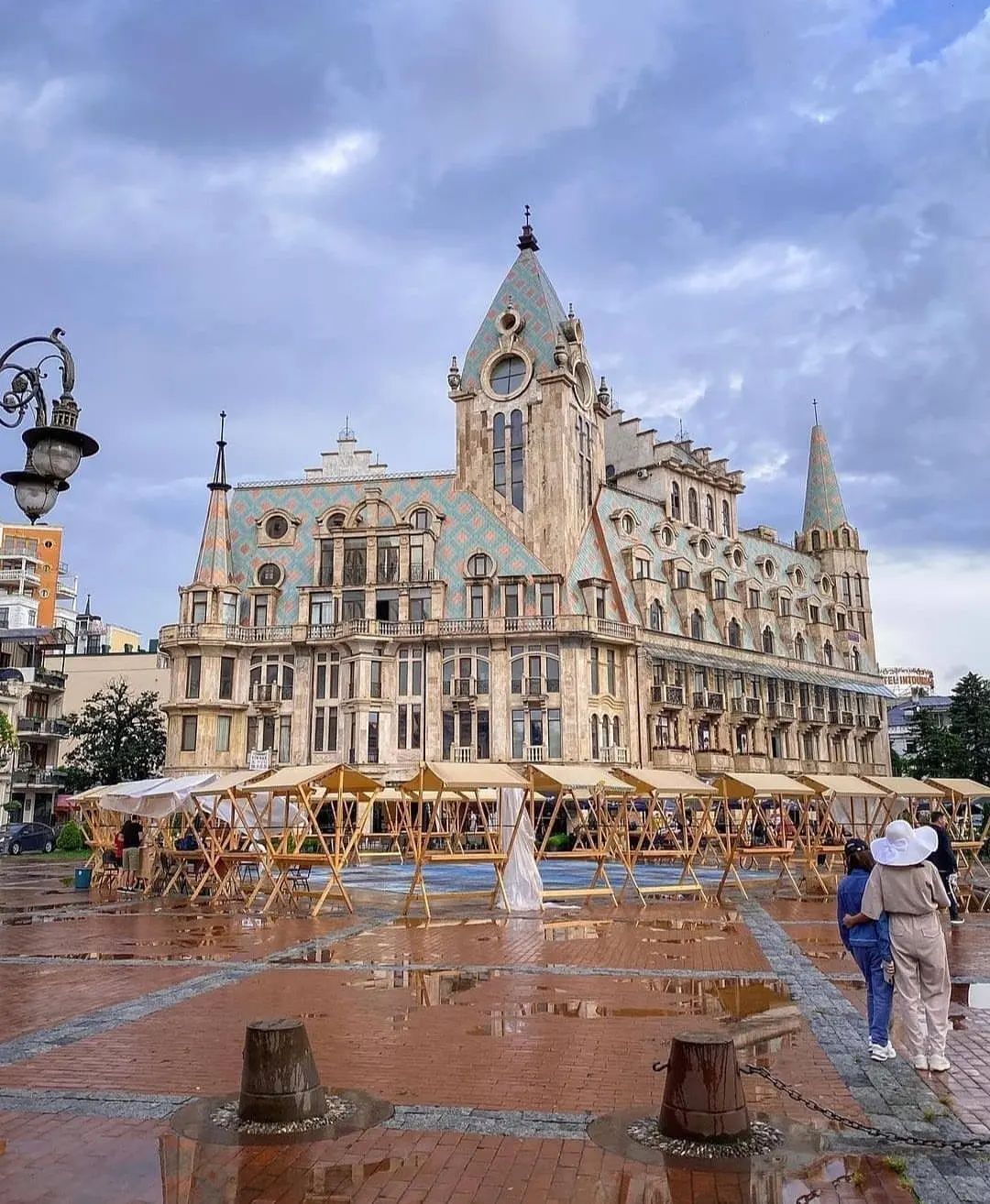 Un edificio histórico con una torre central y múltiples agujas, rodeado por una plaza húmeda con estructuras de madera.