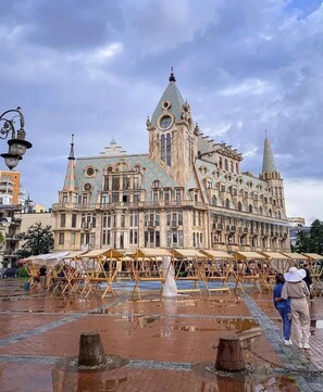 A historic building with a central tower and multiple spires, surrounded by a wet plaza with wooden structures. - Hotel MyRose (Batumi)