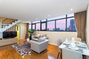 Una sala de estar moderna con una televisión de pantalla plana, un sofá gris, una mesa de comedor para seis personas y un amplio ventanal con vistas a la ciudad.