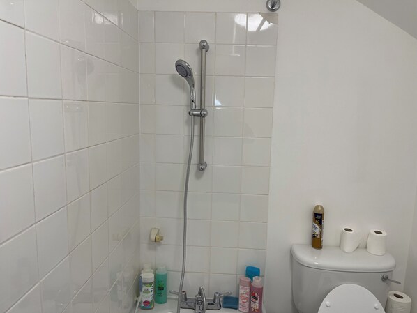 A bathroom with white tiled walls, a shower with a handheld showerhead, a toilet, and various toiletries on the sink.