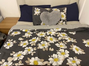 Een bed met een deken met bloemenprint, twee kussens, waarvan één met een hartvormige plaid, en een houten nachtkastje ernaast.