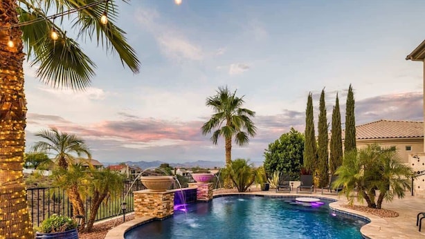 A pool with a waterfall feature, surrounded by palm trees and cypress trees, with a clear sky in the background.