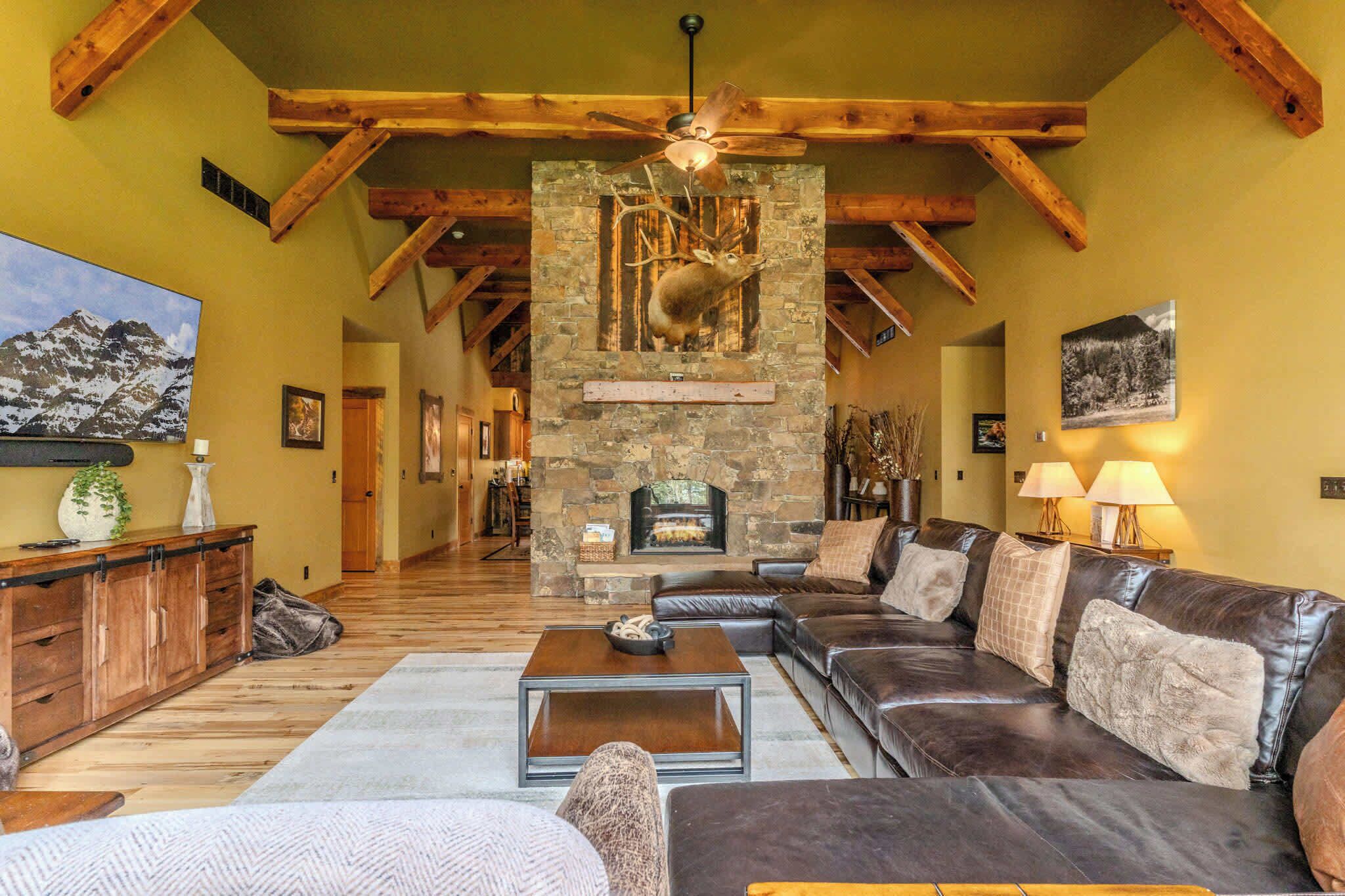 A spacious living room with a stone fireplace, leather sofas, wooden beams, and a mountain painting.