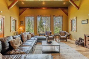 Una sala de estar acogedora con un sofá de cuero, sillones y una mesa de centro. Amplios ventanales ofrecen vistas al bosque.