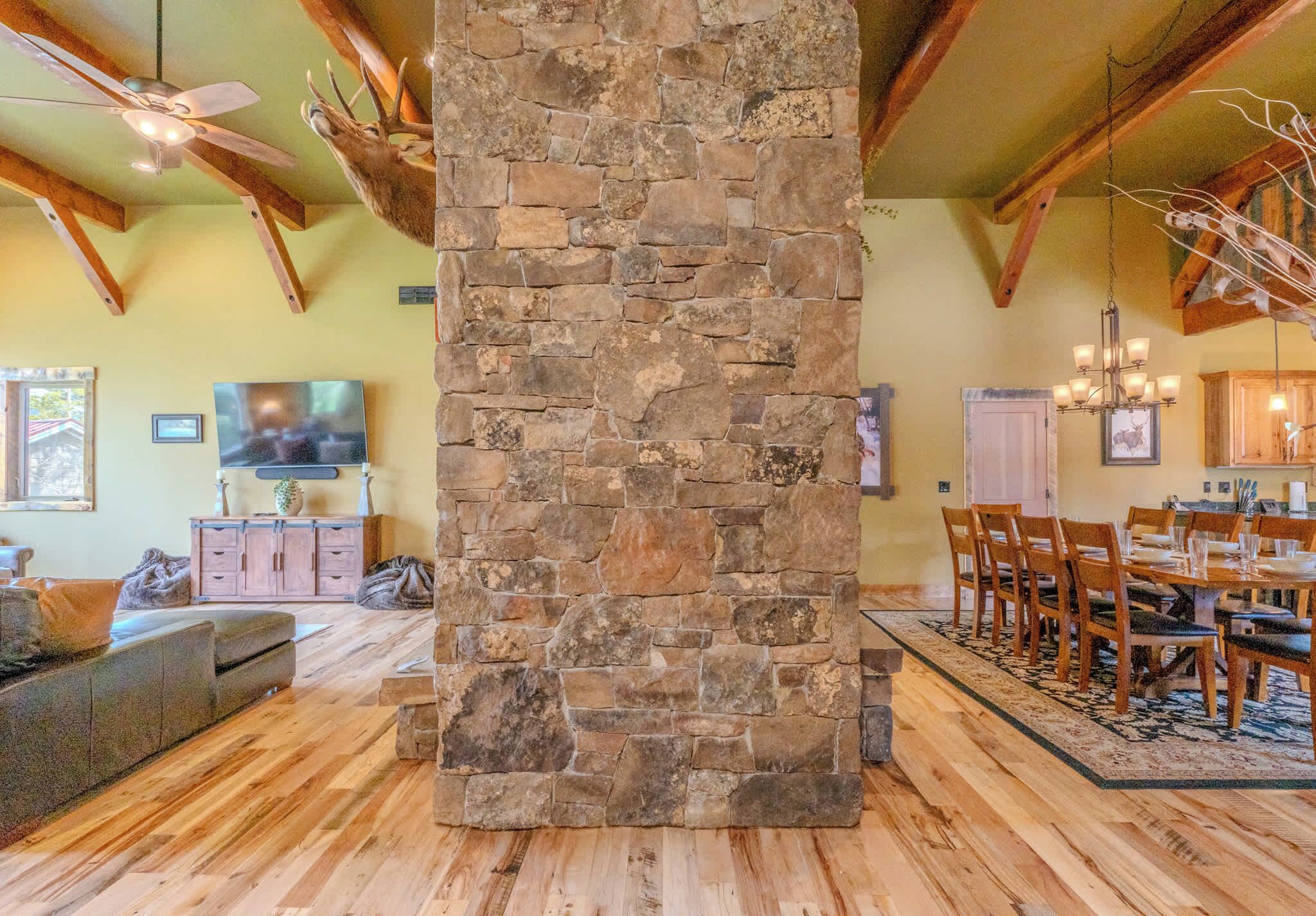 Un pilar de piedra en una habitación con vigas de madera, un televisor, una zona de comedor con sillas y una mesa, y un sofá.
