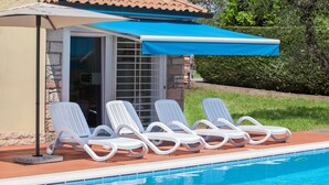 A poolside area with white lounge chairs, a blue awning, and a stone building with glass doors.