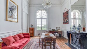 A room with a red sofa, wooden dining table, chandelier, and large windows. - Chateau de Pourpry (Damiatte)