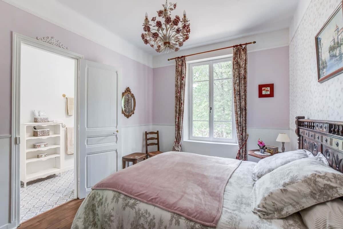 Un dormitorio con cama, una silla, un ventanal con cortinas, una araña y una estantería con objetos.